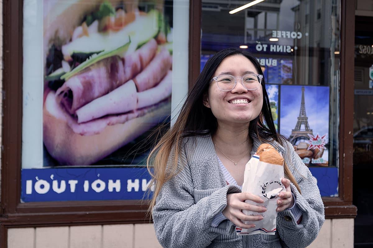 Student holds a banh mi sandwich in front of a shop in Dorchester.